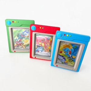 Könnte beinhalten: Drei farbenfrohe Kartenhalter in Grün, Rot und Blau, die jeweils eine Pokémon-Sammelkarte zeigen. Die Halter haben ein Retro-TV-Design mit Bildschirm und Knöpfen. Die Karten zeigen verschiedene Pokémon-Charaktere.