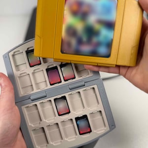 May include: A mustard yellow video game cartridge held above a gray plastic storage case. The case features multiple rectangular slots designed to hold small game cartridges, with several slots currently occupied by black cartridges with red labels.