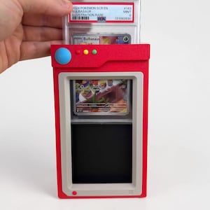 May include: A red and gray plastic display case shaped like a Pokedex, holding a PSA graded 2024 Pokemon Bulbasaur trading card. The case features a blue circular button, three smaller yellow and green buttons, and a rectangular viewing window.