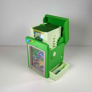 May include: A green plastic trading card deck box with a hinged lid, featuring a front display window showing a card, a top compartment holding a stack of cards, and a small pull-out drawer at the base containing two blue dice for gaming.