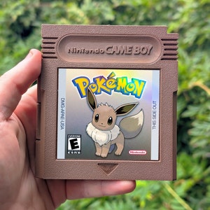 May include: A brown Nintendo Game Boy cartridge featuring the Pokémon game. The label shows the Pokémon logo and an illustration of Eevee. The cartridge has the text "Nintendo GAME BOY" and "THIS SIDE OUT". The game is rated "EVERYONE" by the ESRB.