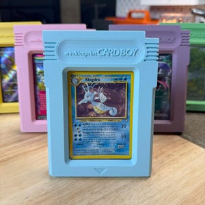 May include: A pastel blue "CARDBOY" case containing a "Kingdra" Pokémon card. The case has the text "Wookieeprint CARDBOY" printed on it. Other coloured cases are visible in the background. The card is a collectible item.