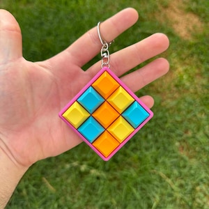 Peut inclure: Un porte-clés coloré de forme carrée avec un cadre rose. L'intérieur est rempli d'une grille de petits carreaux carrés bleus, jaunes et oranges. Le porte-clés est attaché à un anneau en métal argenté.