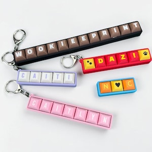 May include: A collection of keychains featuring keyboard-style letter tiles. The keychains are in various colours, including black, red, pink, blue, and purple. Some keychains have a paw print or heart symbol. The words include "WOOKIEEPRINT", "CAITI", "DAZI", "N❤️C", and "CAITLYN".