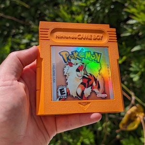 May include: Orange Game Boy cartridge featuring the Pokémon video game. The label displays the Pokémon logo and a fiery creature illustration. The cartridge has the text "Nintendo GAME BOY" and "THIS SIDE OUT".