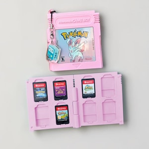 May include: A pink Nintendo Game Boy cartridge case with a Pokemon game cartridge inside. The case is open, revealing slots for Switch game cartridges. A small charm with a cartoon character hangs from the top.