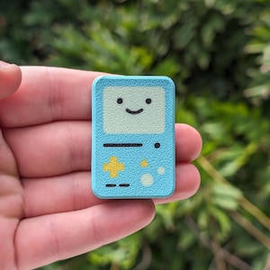 May include: A light blue, square-shaped pin with a smiling face and game controller design. The pin is made of enamel and has a shiny finish.