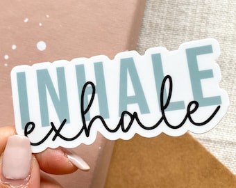 May include: A white sticker with the words "INHALE exhale" in teal and black script. The sticker is held by a person's hand, with a pink background and white speckles.