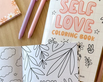 Könnte beinhalten: Ein "Self Love Coloring Book" mit dem Text "Self Love Coloring Book" in Rosa und Orange. Das offene Buch zeigt den Text "You Are Human", umgeben von Strichzeichnungen von Blättern. Rosa und lila Stifte und ein Blumennotizbuch sind ebenfalls zu sehen.
