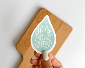 May include: A light blue, teardrop-shaped sticker with the words "DRINK YOUR WATER" in white script. The sticker is held in a hand, with a wooden cutting board in the background. The sticker is a reminder to stay hydrated.