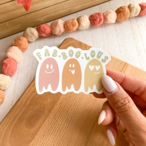 May include: A white sticker with the text "FAB.BOO.LOUS" above three cartoon ghosts. The ghosts are in shades of pink, orange, and yellow, each with a different facial expression. The sticker is on a wooden surface.