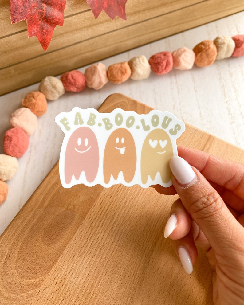 May include: A white sticker with the text "FAB.BOO.LOUS" above three cartoon ghosts. The ghosts are in shades of pink, orange, and yellow, each with a different facial expression. The sticker is on a wooden surface.
