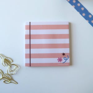 May include: A square notepad featuring horizontal light pink and white stripes with a thin vertical dark line on the left side. The bottom right corner displays a small brown star, a pink flower with a smiling face, and a blue swirling line.