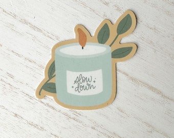 May include: A sticker featuring a light blue candle with a brown flame and the words "slow down" on a white label. The candle is surrounded by green leaves and set against a light brown background.