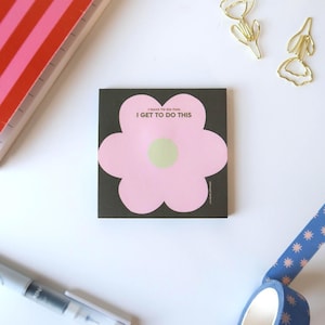 May include: A square sticky note pad with a black background and a large, light pink flower shape in the center. The flower has a pale green circular center and features the text I HAVE TO DO THIS I GET TO DO THIS printed in small dark letters.
