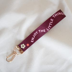 May include: A burgundy wrist strap with the words "ENJOY THE LITTLE THINGS" in white. The strap features a gold-colored clasp and a small floral design. The strap is made of a smooth, satin-like material.