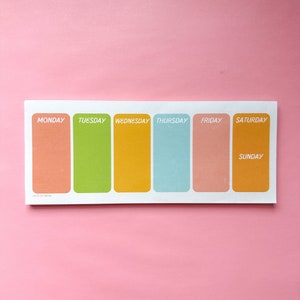 May include: A rectangular weekly planner with a white border and colorful blocks for each day of the week. The days are labeled Monday through Sunday, with each day having a different pastel color. The background is a solid pink.