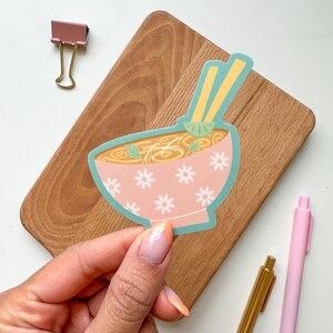 May include: A sticker featuring a pink bowl of ramen with white daisy patterns and a teal border. The bowl contains noodles, broth, and green garnishes, with yellow chopsticks. The sticker is held by a hand, with a wooden cutting board and pens in the background.