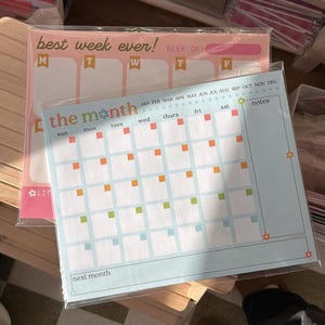 Op de afbeelding: Twee planner pads, één roze en één lichtblauw. De roze pad heeft de tekst "best week ever!" en ruimte voor de week. De blauwe pad toont een maandkalender met de tekst "the month". Perfect voor planning.