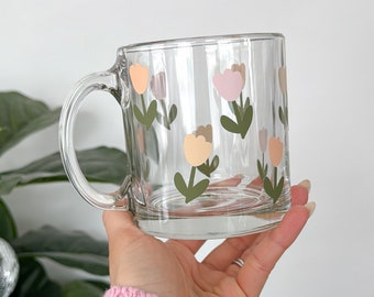 Könnte beinhalten: Transparente Glas-Tasse mit pastellfarbenen Tulpen-Illustrationen. Die Tulpen sind in Pfirsich-, Rosa- und Cremetönen gehalten, mit grünen Stielen und Blättern. Die Tasse hat einen transparenten Henkel und eine quadratische Form.
