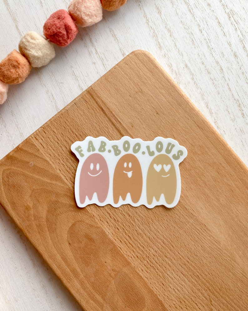 May include: A sticker with the text "FAB.BOO.LOUS" above three smiling ghost illustrations in shades of pink, orange, and yellow. The sticker is on a wooden cutting board. A garland of felt balls in shades of orange and cream is in the background.