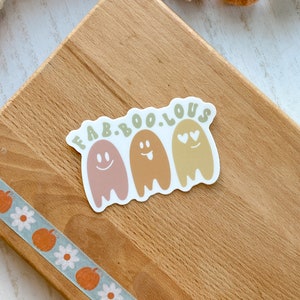 May include: A sticker with the text "FAB.BOO.LOUS" above three cartoon ghosts in shades of pink, brown, and yellow. Each ghost has a different facial expression. A decorative tape with pumpkins and flowers is visible.