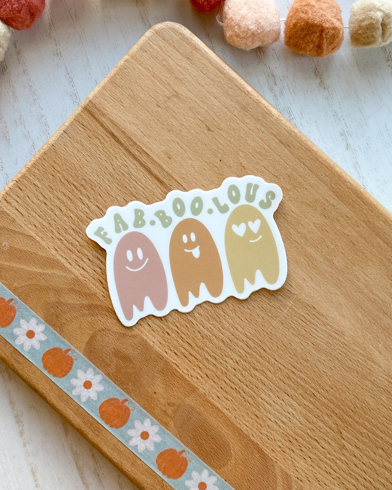 May include: A sticker with the text "FAB.BOO.LOUS" above three cartoon ghosts in shades of pink, brown, and yellow. Each ghost has a different facial expression. A decorative tape with pumpkins and flowers is visible.