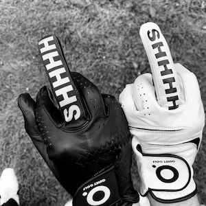 Puede incluir: Un par de guantes de golf negros y blancos con el texto "SHHH" en el dedo índice de cada guante. Los guantes tienen un círculo blanco con el texto "Quiet Golf" en la muñeca.