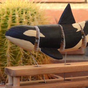 Puede incluir: Una escultura de madera de una orca, pintada de blanco y negro, está suspendida de un soporte de madera con un mecanismo de alambre. El soporte está hecho de madera clara y oscura y tiene una base triangular.