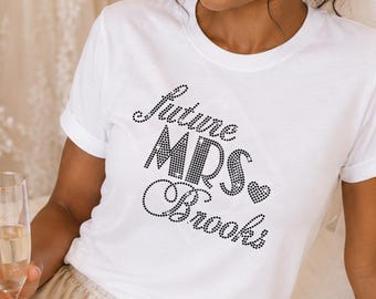 May include: A white short-sleeved t-shirt featuring the text "Future MRS Brooks" in black rhinestones, paired with champagne-colored satin shorts. The shirt has a crew neckline and a fitted silhouette, while the shorts have a drawstring waist.