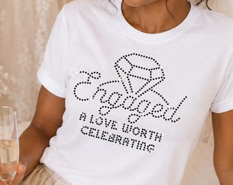 May include: A white short-sleeved t-shirt featuring a black rhinestone graphic of a diamond and the text "Engaged A LOVE WORTH CELEBRATING" paired with champagne-colored satin shorts with a drawstring waist and ruffled hemline.