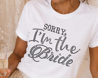 May include: A white short-sleeved t-shirt featuring the text "SORRY, I'm the Bride" in black rhinestones. The shirt is paired with champagne-colored satin shorts with a drawstring waist. A sheer white veil is attached to the hair.