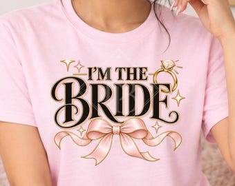 May include: A light pink short-sleeve t-shirt featuring the text I'M THE BRIDE in black lettering, accompanied by a graphic of a gold engagement ring and a pink ribbon bow. The shirt is displayed as a front view mockup for design purposes.