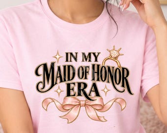 May include: A light pink t-shirt featuring a graphic print with the text "IN MY MAID OF HONOR ERA" in black lettering, accompanied by a gold diamond ring illustration and a pink ribbon design centered on the chest of the garment.