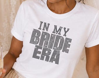 May include: A white short-sleeved t-shirt featuring the black text IN MY BRIDE ERA in a studded or rhinestone-style font. The shirt is paired with light champagne-colored satin shorts with a drawstring waist and a ruffled hemline.
