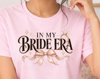 May include: A light pink short-sleeve t-shirt featuring the text IN MY BRIDE ERA in black lettering, accompanied by a graphic of a diamond ring and a ribbon bow. The shirt is displayed as a front view mockup with additional text overlays.