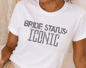May include: A white short-sleeved t-shirt featuring the text BRIDE STATUS: ICONIC in black rhinestones, paired with champagne-colored satin shorts. The shirt has a crew neckline and a standard fit, while the shorts feature an elasticized waistband.