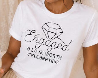 May include: A white short-sleeved t-shirt featuring a black rhinestone graphic of a diamond and the text "Engaged A LOVE WORTH CELEBRATING" paired with champagne-colored satin shorts with a drawstring waist and ruffled hemline.
