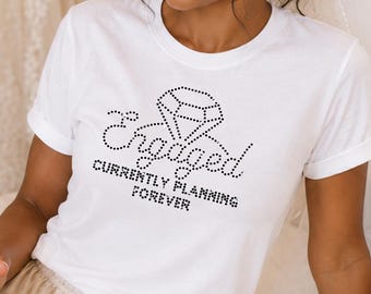 May include: A white short-sleeved t-shirt featuring a black rhinestone graphic of a diamond and the text Engaged CURRENTLY PLANNING FOREVER. The shirt is paired with champagne-colored satin shorts with a drawstring waist and ruffled hem.