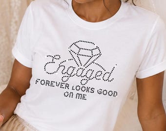 May include: A white short-sleeved t-shirt featuring a rhinestone graphic of a diamond ring and the text Engaged FOREVER LOOKS GOOD ON ME. The shirt is paired with champagne-colored satin shorts with a drawstring waist and a white bridal veil.