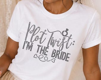 May include: A white short-sleeved t-shirt featuring black rhinestone text that reads Plot Twist: I'M THE BRIDE with a small ring icon. The shirt is paired with champagne-colored satin shorts and a sheer white bridal veil with a beaded edge.
