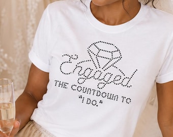 May include: A white short-sleeved t-shirt featuring a black rhinestone graphic of a diamond ring and the text "Engaged THE COUNTDOWN TO "I DO."" paired with champagne-colored satin shorts with a drawstring waist and a sheer white veil.