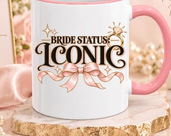 May include: An 11 oz white ceramic mug with a pink handle and interior rim. The front features the text BRIDE STATUS: ICONIC in black, a gold diamond ring graphic, and a pink ribbon illustration. The mug sits on a textured stone coaster.