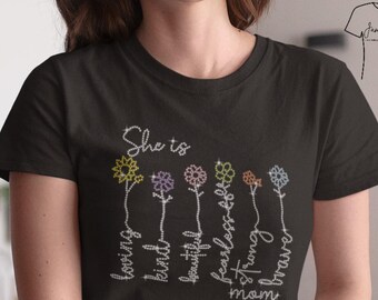 以下が含まれることがあります： 「She is」の文字とカラフルな花のイラストがプリントされた黒いTシャツ。「loving, kind, beautiful, fearless, strong, brave, mom」の文字が白い筆記体で書かれています。