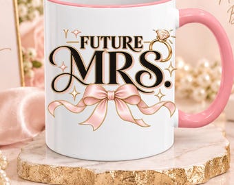 May include: A white ceramic mug with a pink handle and rim, featuring the black text 'FUTURE MRS.' above a pink ribbon illustration and a gold diamond ring graphic. The mug is labeled as an 11 oz pink accent mug with design placement options.