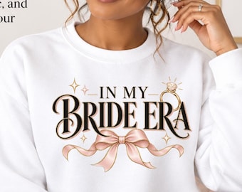 May include: A white crewneck sweatshirt featuring a graphic print that reads "IN MY BRIDE ERA" in black lettering, accompanied by a gold engagement ring illustration and a light pink ribbon bow, with small star accents surrounding the text.