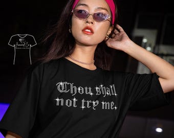 以下が含まれることがあります： ラインストーンのようなフォントで「Thou shall not try me」と書かれた黒いTシャツ。モデルはピンクのヘアバンド、サングラスを着用し、赤いハンドバッグを持っています。このシャツはアパレルコレクションの一部です。