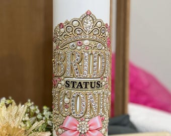 May include: A white cylindrical tumbler featuring a gold-toned crown graphic with embedded rhinestones, the text "BRIDE STATUS: ICONIC" in gold lettering, and a pink bow accent at the base. A clear plastic straw is inserted into the lid of the tumbler.