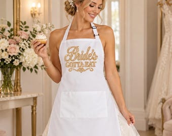 May include: A white apron featuring a large front pocket and the gold glitter text "Bride's GOTTA EAT" printed on the chest. The apron has a halter-style neck strap and is worn over a white lace gown with a full skirt and a sweetheart neckline.