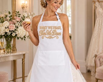 May include: A white apron featuring a front pocket and gold glitter text that reads First We Feast, Then We Dance. The apron has a halter-style neck strap and ties at the waist, designed to be worn over a full-length white lace wedding gown.
